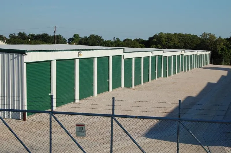Rows of mini-storage units with green roll-up doors behind security fencing.